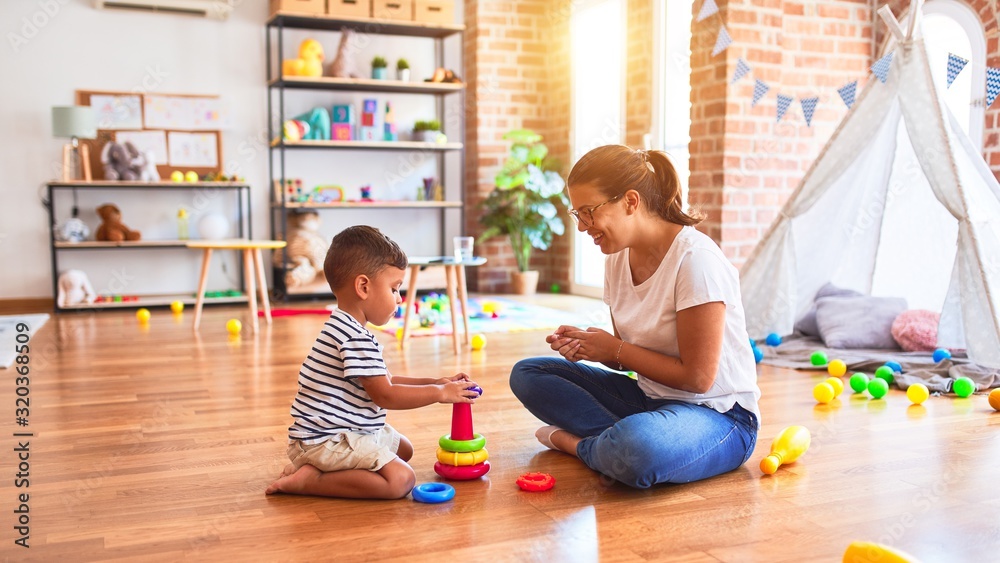 Fototapeta premium Beautiful teacher and toddler boy building pyramid with hoops bolcks at kindergarten