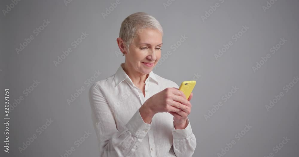 Elderly woman using smartphone and smiling on gray background