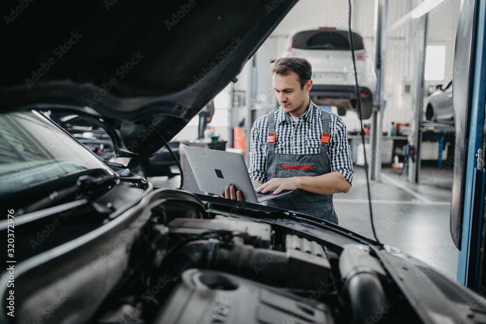 Smiling young man mechanic using a laptop computer to check a car engine Smiling young man mechanic using a laptop computer to check a car engine