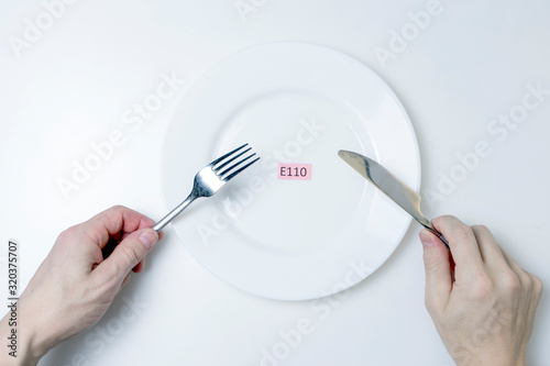 Harmful food additives. Men's hands hold a knife and a fork. On the plate is a plate with the E-additive code.