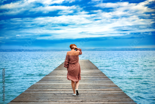 Photography Back of faceless red haired woman in red dress walking by wooden pier on Garda lake in cloudy day