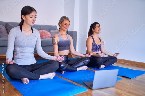 Young beautiful group of sportswomen practicing yoga. Doing lotus pose using laptop at gym