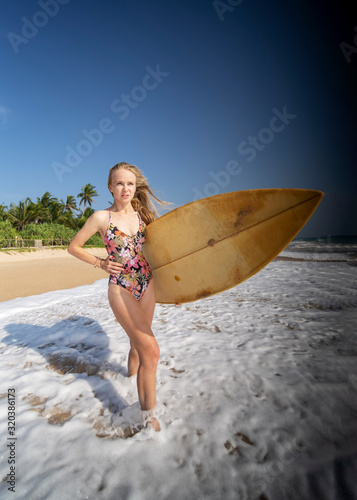 A young woman walks along the beach with a surfboard on the background of the ocean and the sun