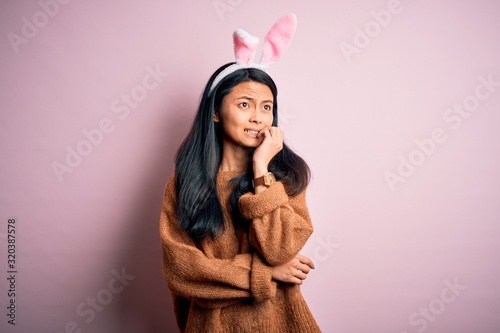 Young beautiful chinese woman wearing bunny ears standing over isolated pink background looking stressed and nervous with hands on mouth biting nails. Anxiety problem.