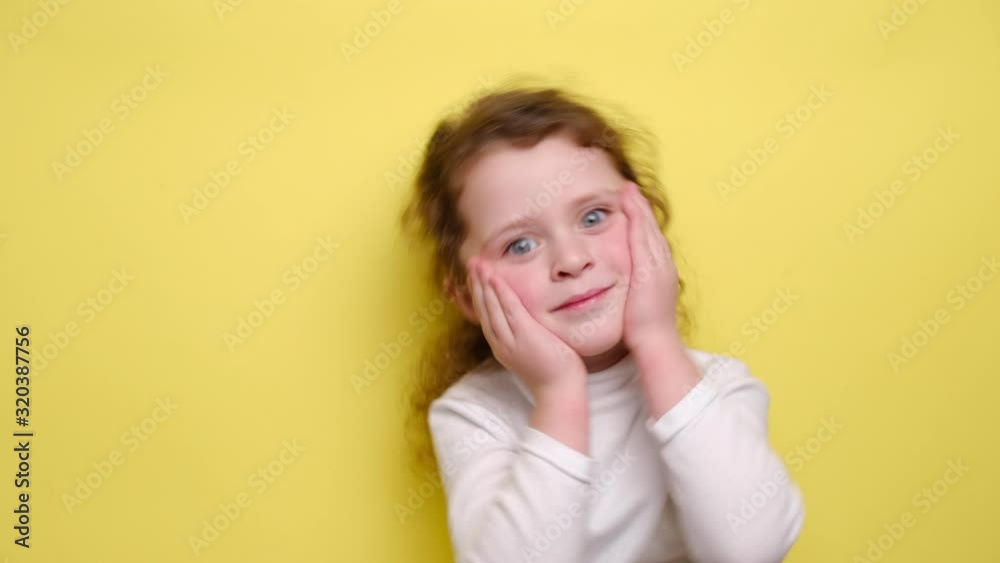 Happy child smiles keeps hand near cheek, romantic expression, remembers something touching or pleasant, wears white jumper, looking at camera, standing over yellow background. Happy child smiles keeps hand near cheek, romantic expression, remembers something touching or pleasant, wears white jumper, looking at camera, standing over yellow background.