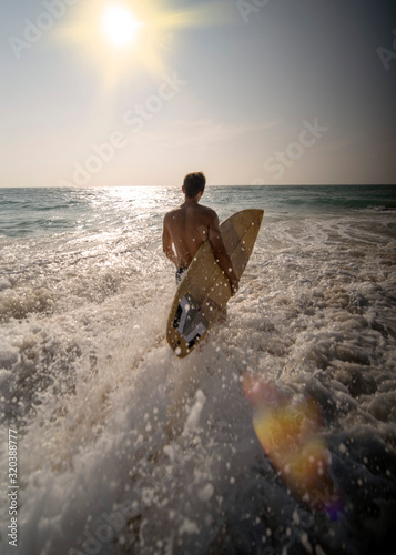 Athletic male surfer, goes with a surfboard into the ocean on the oncoming sun and waves.