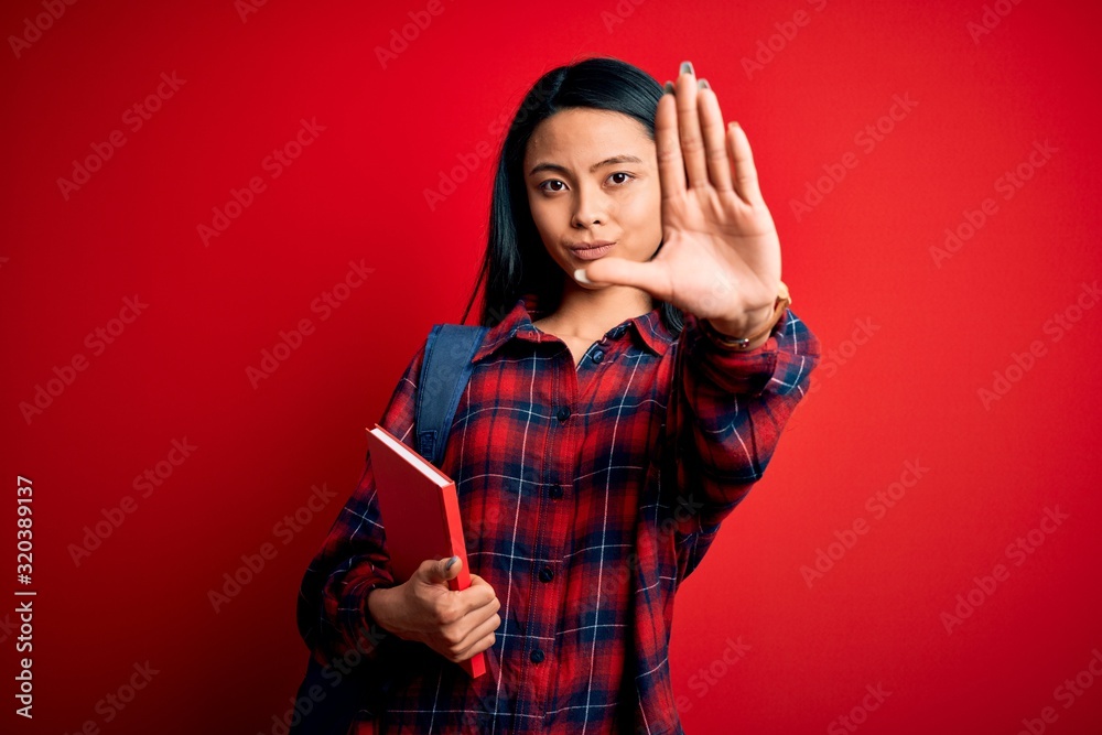 Naklejka premium Young beautiful chinese student woman holding book standing over isolated pink background with open hand doing stop sign with serious and confident expression, defense gesture