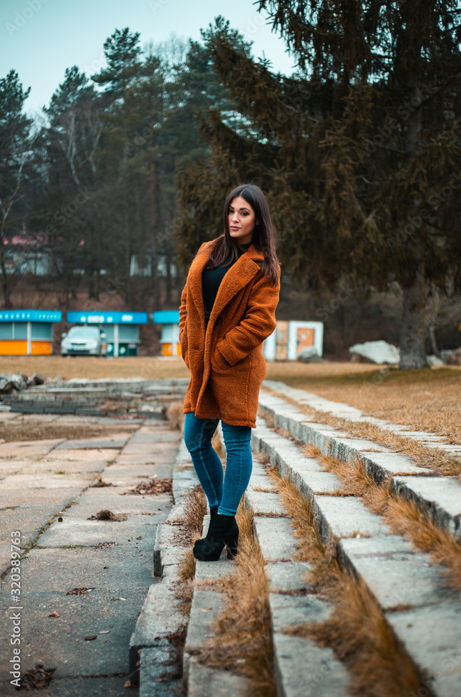 Portrait of beautiful brunette girl (woman) in orange fashion coat in season - photo on stairs with park and nature on background. Young female model posing and looking to camera - full body shot. Portrait of beautiful brunette girl (woman) in orange fashion coat in season - photo on stairs with park and nature on background. Young female model posing and looking to camera - full body shot.