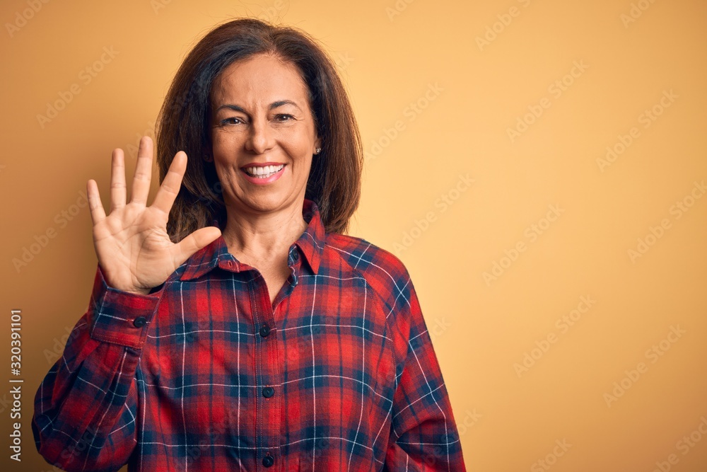 Middle age beautiful woman wearing casual shirt standing over isolated yellow background showing and pointing up with fingers number five while smiling confident and happy.