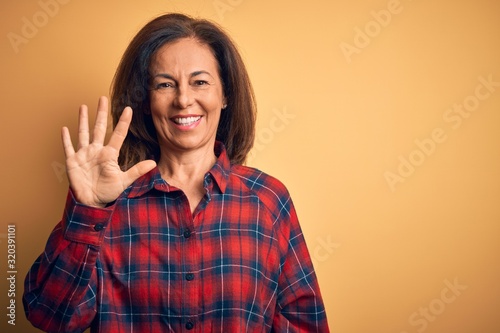 Middle age beautiful woman wearing casual shirt standing over isolated yellow background showing and pointing up with fingers number five while smiling confident and happy.