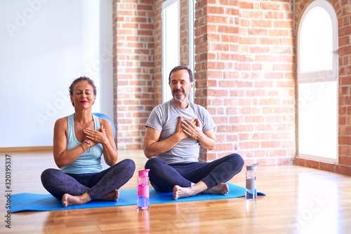 Middle age sporty couple sitting on mat doing stretching yoga exercise at gym smiling with hands on chest with closed eyes and grateful gesture on face. Health concept.