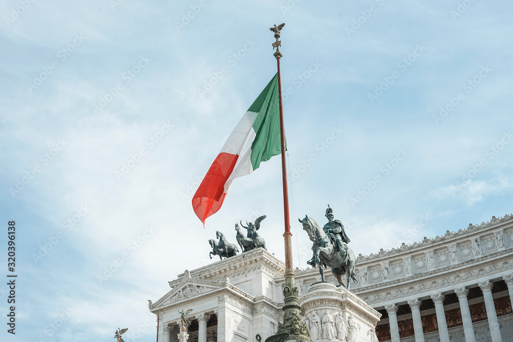 View of Italian national flag in front of Altare della Patria (Altar of the Fatherland) , the equestrian sculpture of Victor Emmanuel and statue of the goddess Victoria riding on quadrigas on top.