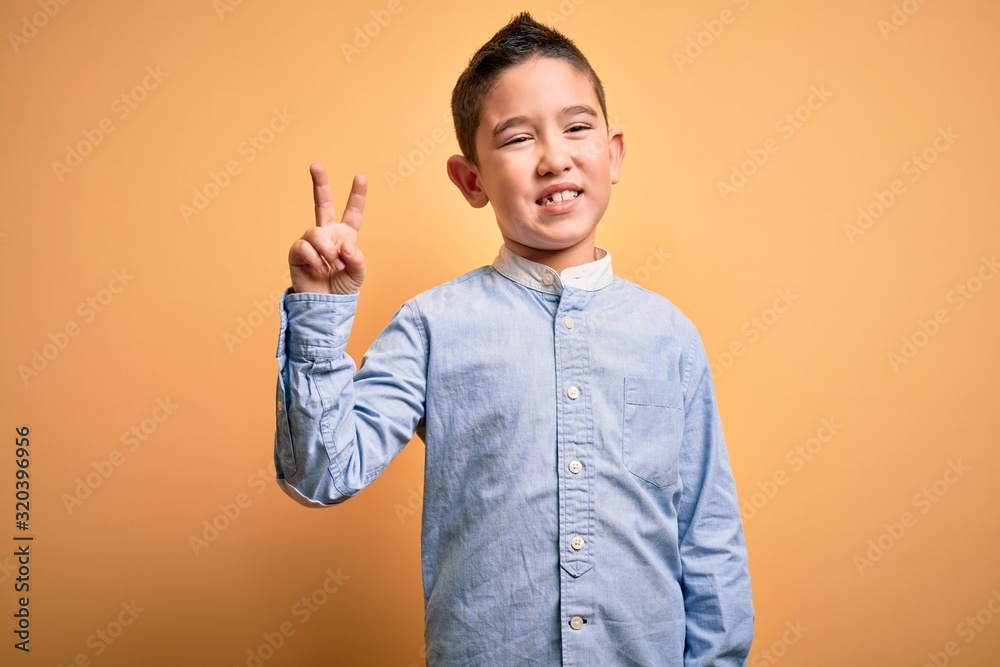 Young little boy kid wearing elegant shirt standing over yellow isolated background smiling with happy face winking at the camera doing victory sign. Number two. Young little boy kid wearing elegant shirt standing over yellow isolated background smiling with happy face winking at the camera doing victory sign. Number two.