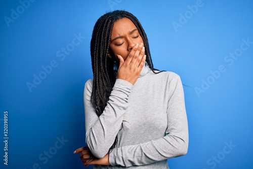 Young african american woman standing wearing casual turtleneck over blue isolated background bored yawning tired covering mouth with hand. Restless and sleepiness.