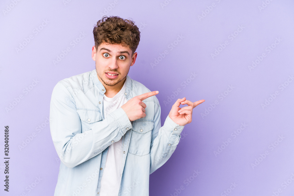 Young blond curly hair caucasian man isolated shocked pointing with index fingers to a copy space. Young blond curly hair caucasian man isolated shocked pointing with index fingers to a copy space.