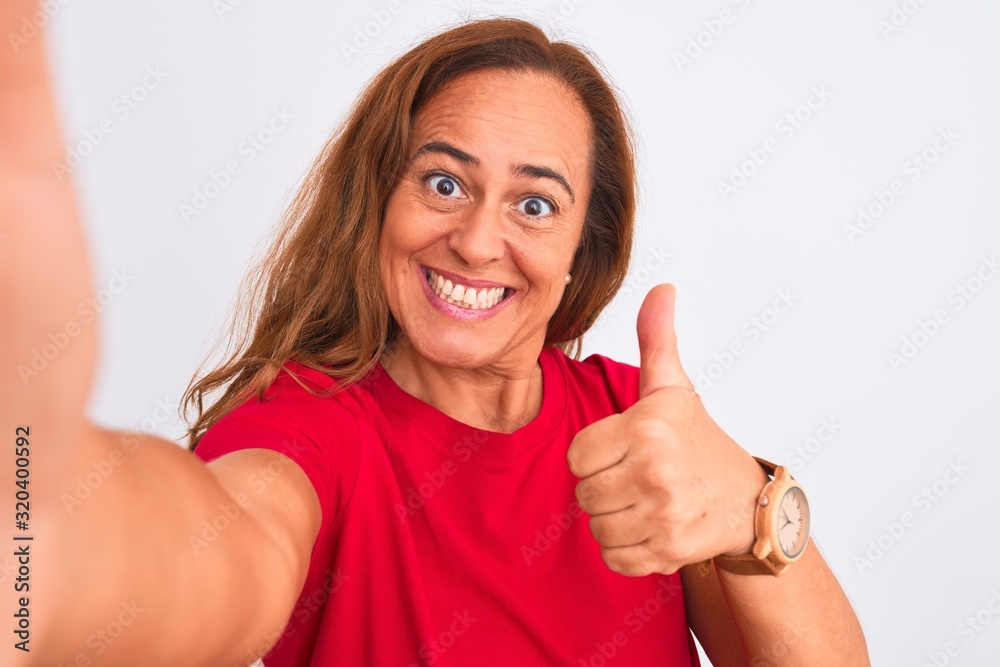 Middle age mature woman taking a selfie photo using smartphone over isolated background happy with big smile doing ok sign, thumb up with fingers, excellent sign Middle age mature woman taking a selfie photo using smartphone over isolated background happy with big smile doing ok sign, thumb up with fingers, excellent sign