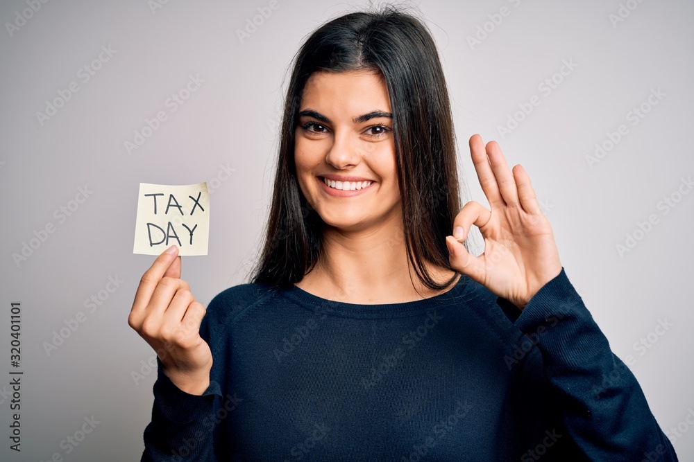 Young beautiful brunette woman holding paper with reminder paper with tax day message doing ok sign with fingers, excellent symbol