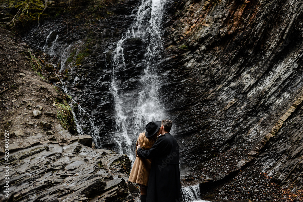 a couple in love kisses near a waterfall, a man is dressed in a stylish coat, a hat on the girl head. Stylish guys travel the world.