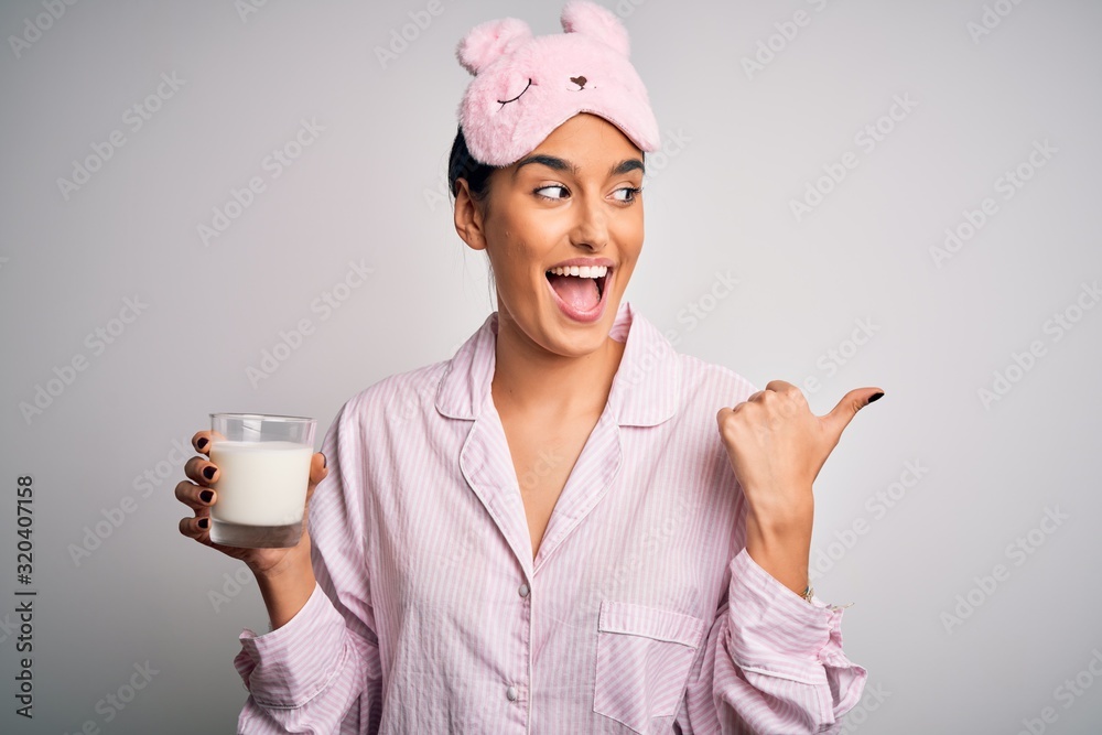 Young beautiful woman wearing pajama and sleep mask drinking glass of healthy milk pointing and showing with thumb up to the side with happy face smiling Young beautiful woman wearing pajama and sleep mask drinking glass of healthy milk pointing and showing with thumb up to the side with happy face smiling