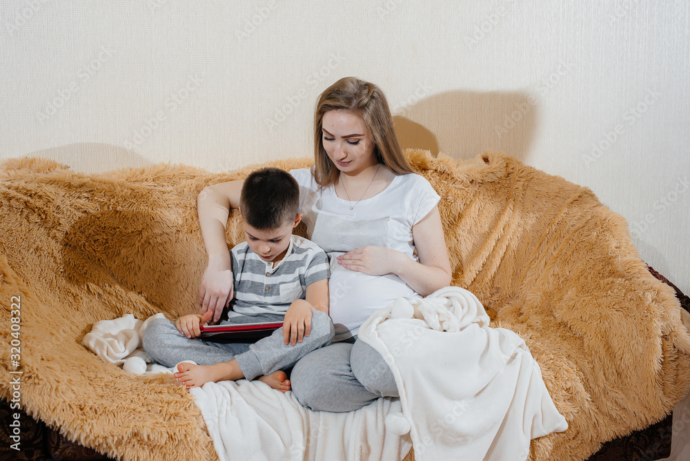 Pregnant mom plays with her son on the couch in the tablet. Care, family, love Pregnant mom plays with her son on the couch in the tablet. Care, family, love
