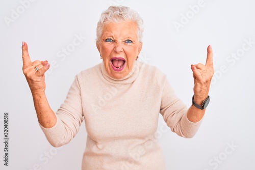 Senior grey-haired woman wearing turtleneck sweater standing over isolated white background shouting with crazy expression doing rock symbol with hands up. Music star. Heavy music concept.
