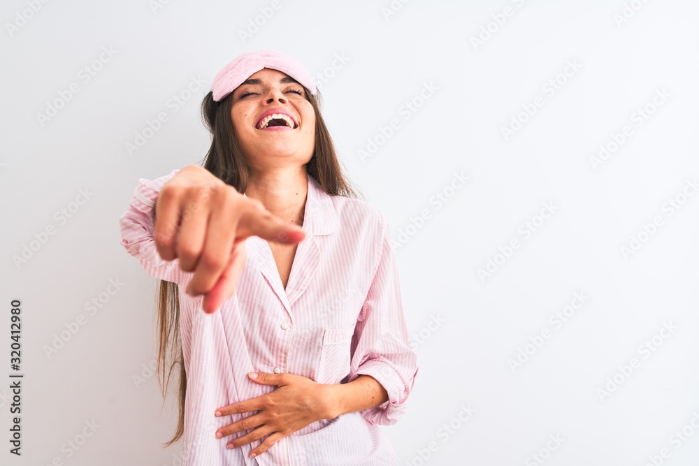 Young beautiful woman wearing sleep mask and pajama over isolated white background laughing at you, pointing finger to the camera with hand over body, shame expression Young beautiful woman wearing sleep mask and pajama over isolated white background laughing at you, pointing finger to the camera with hand over body, shame expression
