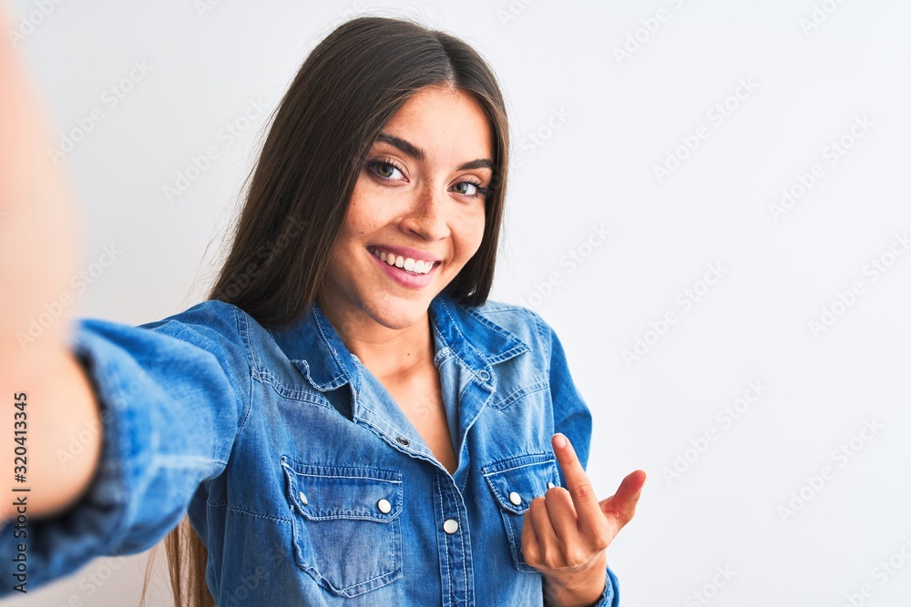 Beautiful woman wearing denim shirt make selfie by camera over isolated white background Beckoning come here gesture with hand inviting welcoming happy and smiling Beautiful woman wearing denim shirt make selfie by camera over isolated white background Beckoning come here gesture with hand inviting welcoming happy and smiling