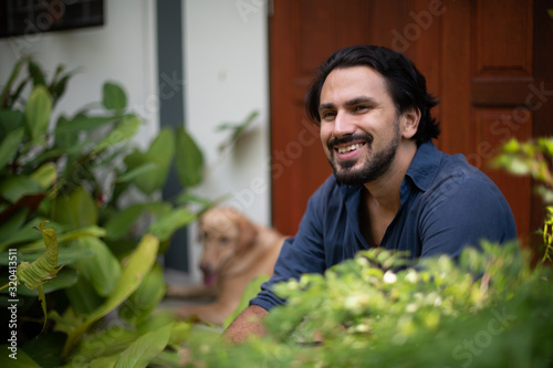 A man with dogs sits on the porch of a house in a tropical garden. © Anna