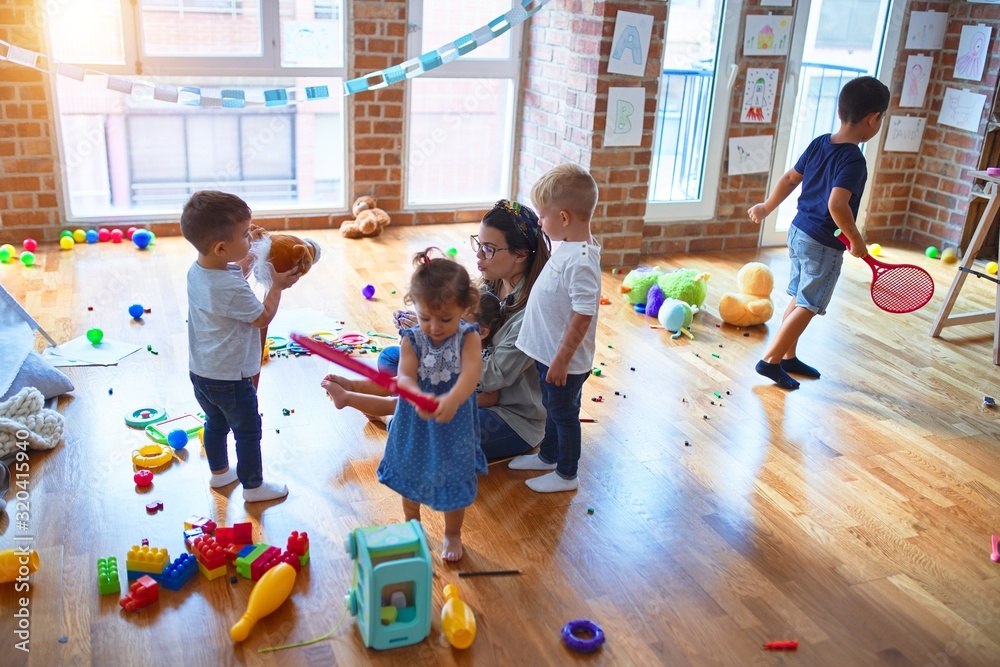 Beautiful teacher and group of toddlers playing around lots of toys at kindergarten - obrazy, fototapety, plakaty 