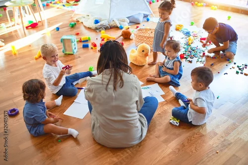 Obraz Beautiful teacher and group of toddlers sitting on the floor drawing using paper and pencil around lots of toys at kindergarten