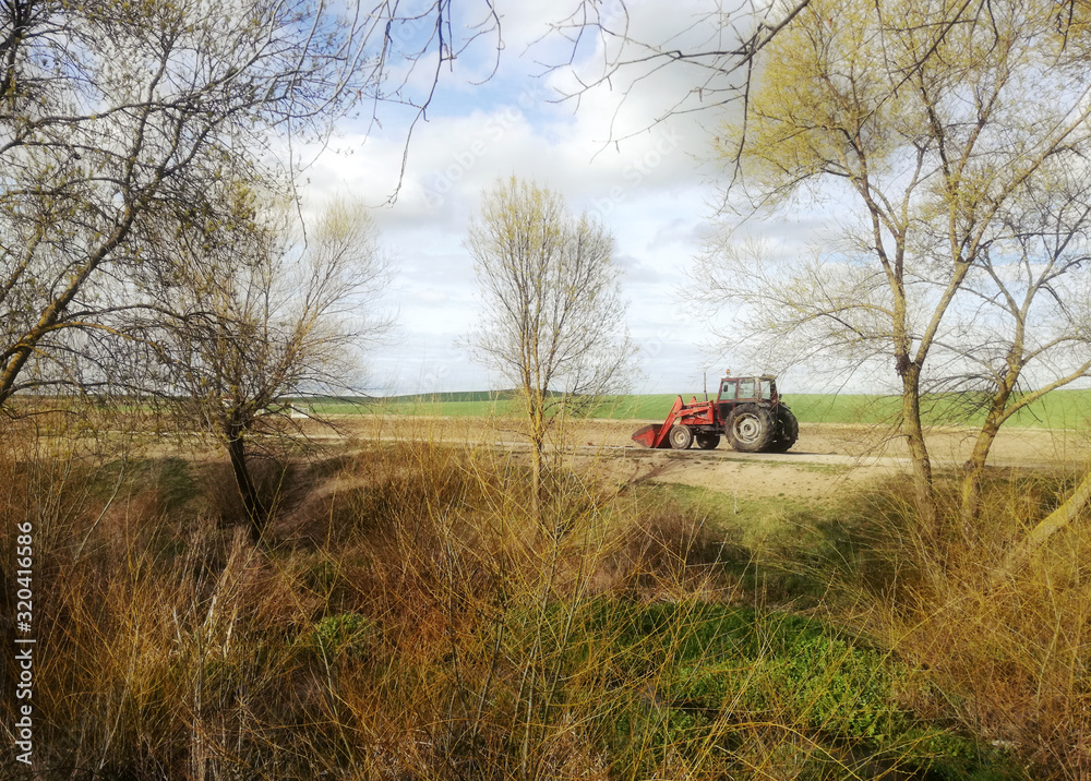 Fototapeta premium Tractor with shovel on the dirt road next to the river. Genuine print of agricultural area. Rural environment. Spanish field Landscape with crops. Horizon in day with calm sky. Trees and low bushes.