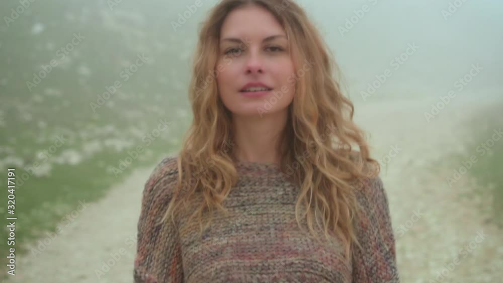 Portrait curly woman walk in fog, mountain road way at peak top of Monte Baldo. beckoning calling gesture follow me. Smiling cute attractive face with long flowing blond hair. Knitted warm sweater Portrait curly woman walk in fog, mountain road way at peak top of Monte Baldo. beckoning calling gesture follow me. Smiling cute attractive face with long flowing blond hair. Knitted warm sweater