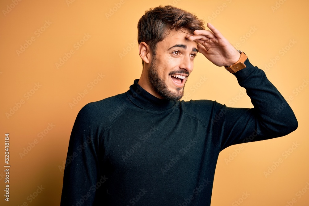 Young handsome man with beard wearing turtleneck sweater standing over yellow background very happy and smiling looking far away with hand over head. Searching concept. Young handsome man with beard wearing turtleneck sweater standing over yellow background very happy and smiling looking far away with hand over head. Searching concept.