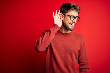 Young handsome man with beard wearing glasses and sweater standing over red background smiling with hand over ear listening an hearing to rumor or gossip. Deafness concept.