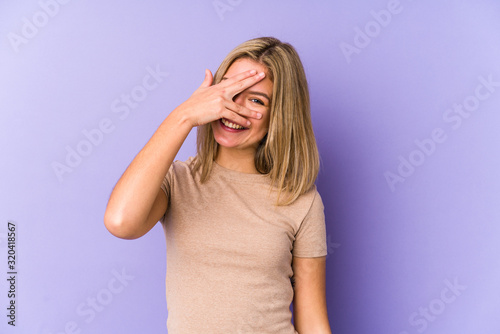 Young blonde caucasian woman isolated blink at the camera through fingers, embarrassed covering face.