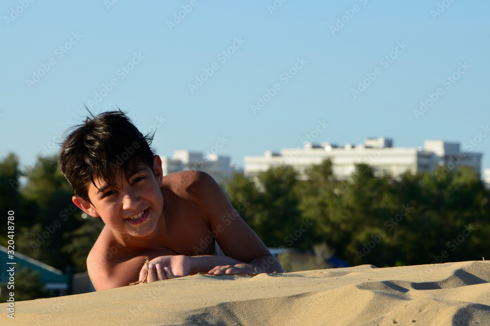 A dark-haired boy in swimming trunks is lying on the yellow sand on a Sunny day and smiling at the camera. Summer vacation on the beach. Selective focus. A dark-haired boy in swimming trunks is lying on the yellow sand on a Sunny day and smiling at the camera. Summer vacation on the beach. Selective focus.