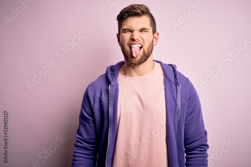 Young blond man with beard and blue eyes wearing purple sweatshirt over pink background sticking tongue out happy with funny expression. Emotion concept.