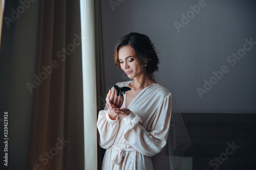 A beautiful gentle bride in a beige peignoir with a butterfly with black and blue wings sits on the hand of the bride, she smiles