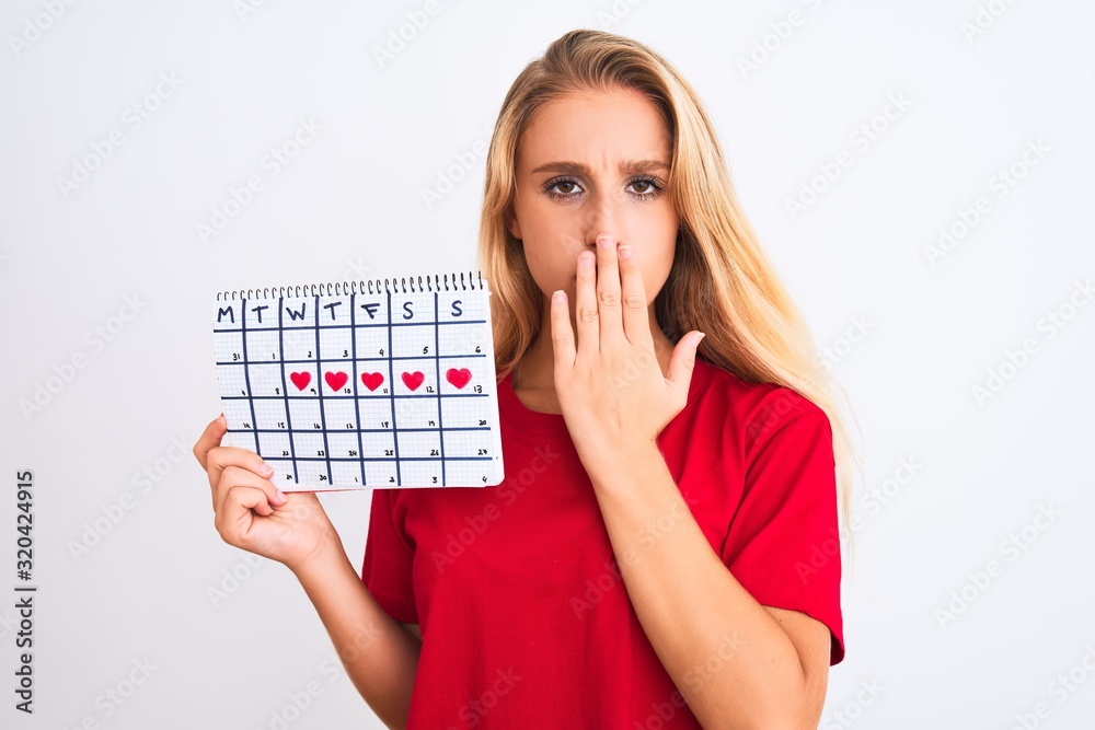 Young beautiful woman holding period calendar standing over isolated white background cover mouth with hand shocked with shame for mistake, expression of fear, scared in silence, secret concept
