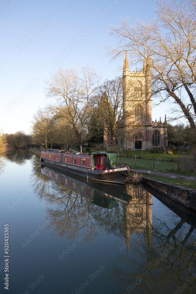 A peaceful winter’s afternoon with a narrowboat and St Lawrences Church, Hungerford reflected in the still water of the Kennet and Avon Canal, Berkshire, UK