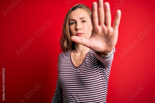 Young beautiful blonde woman wearing casual striped t-shirt over isolated red background doing stop sing with palm of the hand. Warning expression with negative and serious gesture on the face.