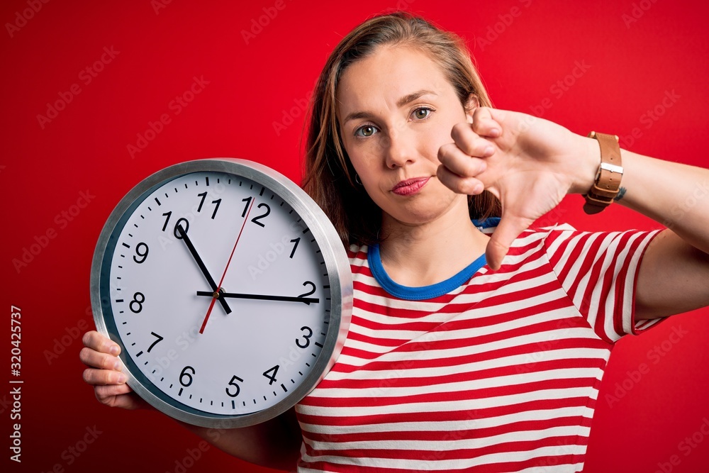 Young beautiful blonde woman holding big clock standing over isolated red background with angry face, negative sign showing dislike with thumbs down, rejection concept Young beautiful blonde woman holding big clock standing over isolated red background with angry face, negative sign showing dislike with thumbs down, rejection concept