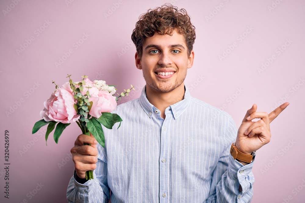 Young blond man with curly hair holding beautiful bouquet over isolated pink background very happy pointing with hand and finger to the side Young blond man with curly hair holding beautiful bouquet over isolated pink background very happy pointing with hand and finger to the side