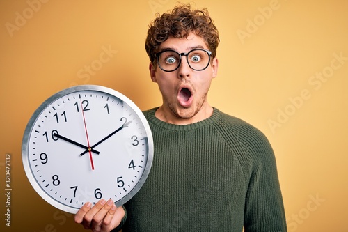 Young blond man with curly hair wearing glasses holding big clock over yellow background scared in shock with a surprise face, afraid and excited with fear expression photo