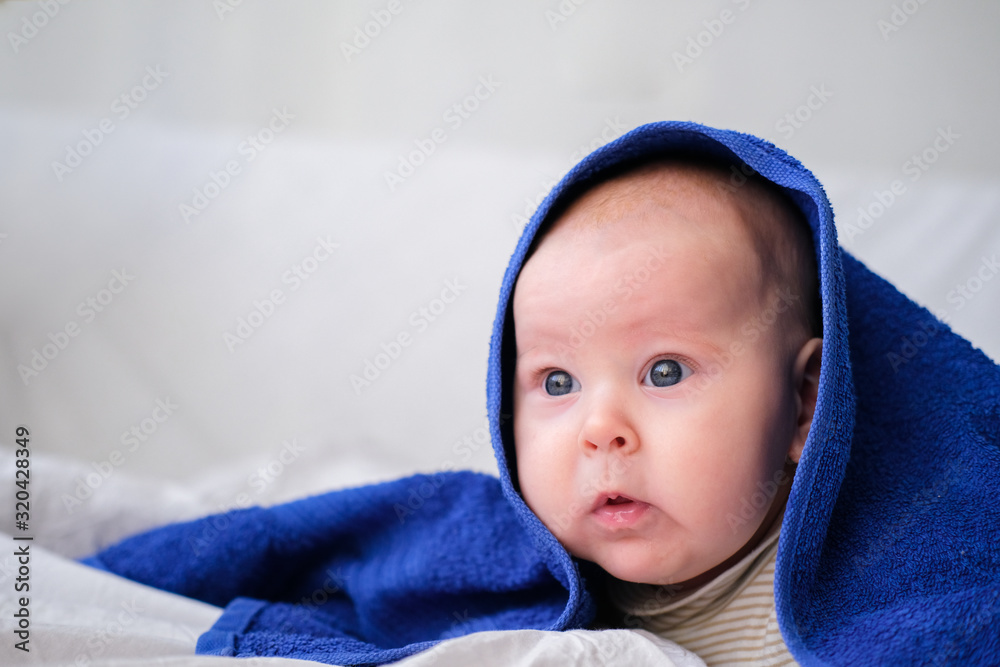 Newborn caucasian infant baby wrapped in a blue towel looks puzzled to the side after bathing in the bath on white bed. Copyspace Newborn caucasian infant baby wrapped in a blue towel looks puzzled to the side after bathing in the bath on white bed. Copyspace
