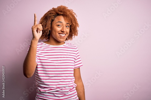 Young beautiful African American afro woman with curly hair wearing casual striped t-shirt pointing finger up with successful idea. Exited and happy. Number one.