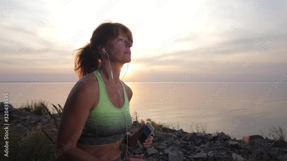 Close-up portrait of positive sporty redhead woman listening to music on phone while jogging along sea shore at sunset. Active mature runner in headphones enjoying music and cardio training in nature Close-up portrait of positive sporty redhead woman listening to music on phone while jogging along sea shore at sunset. Active mature runner in headphones enjoying music and cardio training in nature