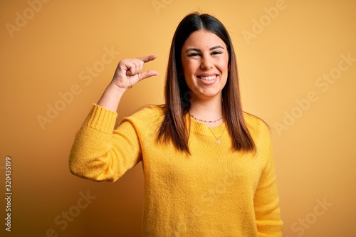 Young beautiful woman wearing casual sweater over yellow isolated background smiling and confident gesturing with hand doing small size sign with fingers looking and the camera. Measure concept.