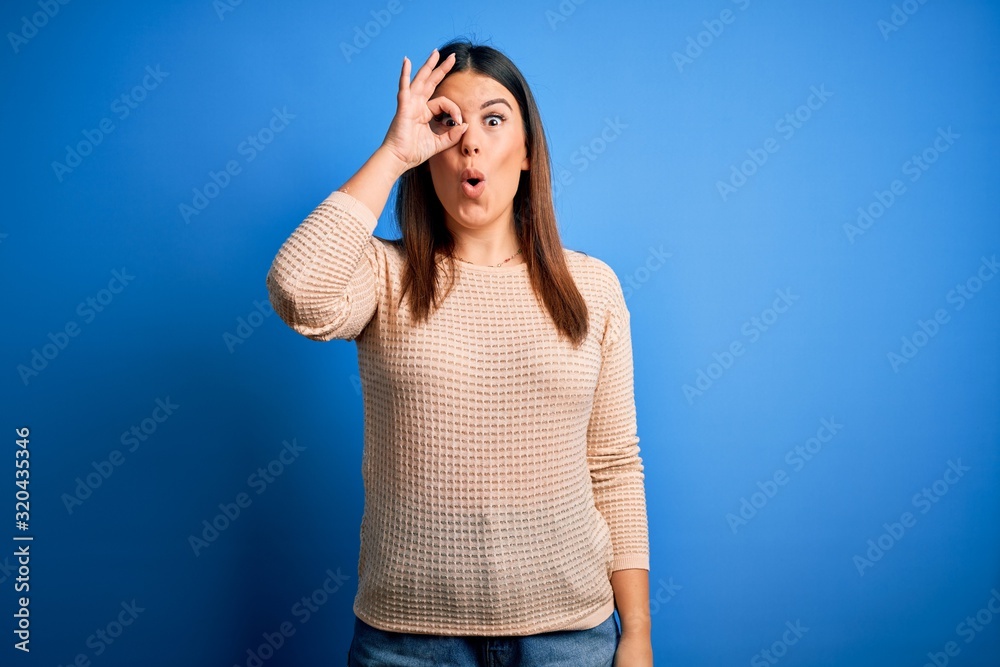 Young beautiful woman wearing casual sweater over blue background doing ok gesture shocked with surprised face, eye looking through fingers. Unbelieving expression. Young beautiful woman wearing casual sweater over blue background doing ok gesture shocked with surprised face, eye looking through fingers. Unbelieving expression.