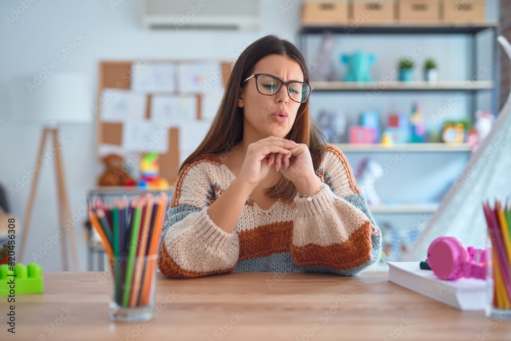 Young beautiful teacher woman wearing sweater and glasses sitting on desk at kindergarten Suffering pain on hands and fingers, arthritis inflammation